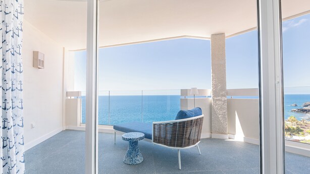 Un balcón con vista al mar, una silla de mimbre, una mesita redonda y una cortina estampada.