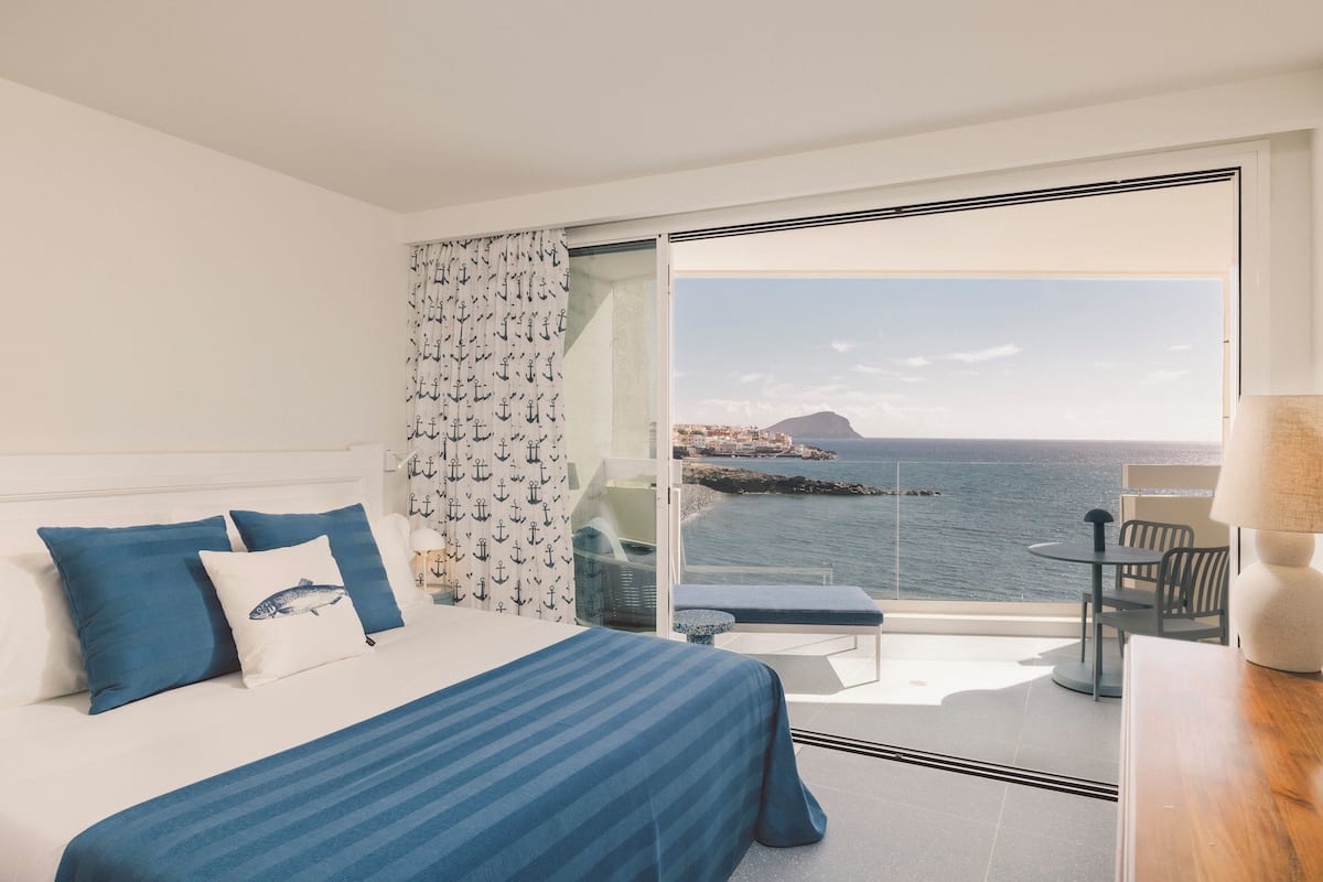 A bedroom with a bed, a bench, and a table with a lamp. There is a view of the sea and a distant island through a glass door.
