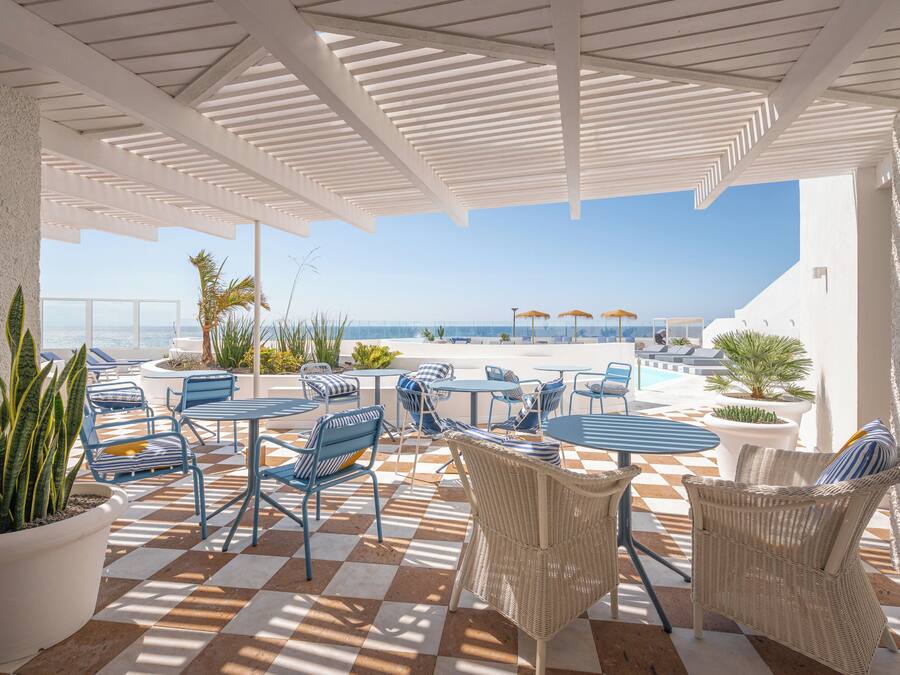 A covered outdoor seating area with checkered flooring, blue and wicker furniture, and a view of the sea.