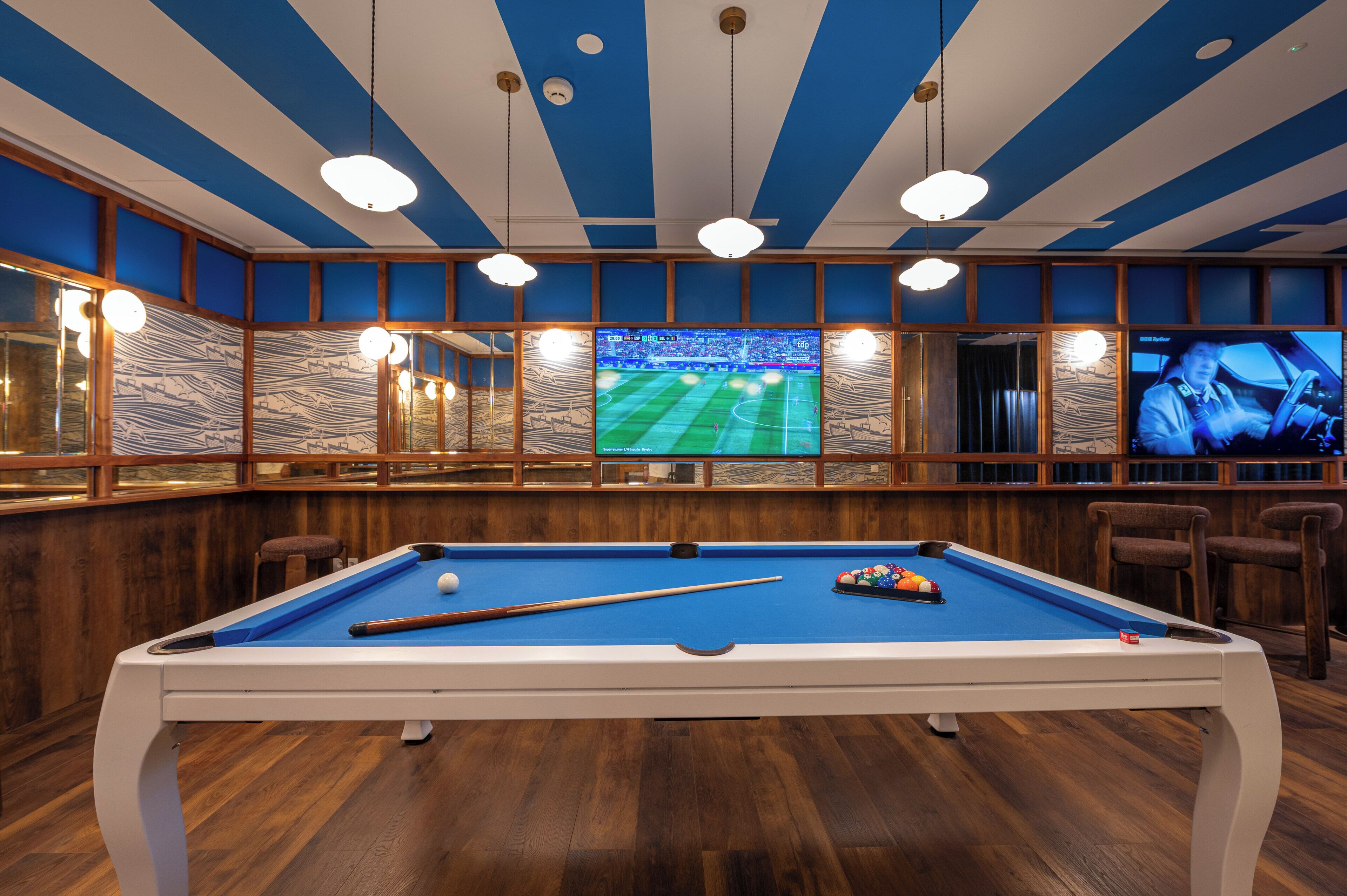 A pool table with a blue felt surface, surrounded by wooden paneling and a large screen displaying a sports event.