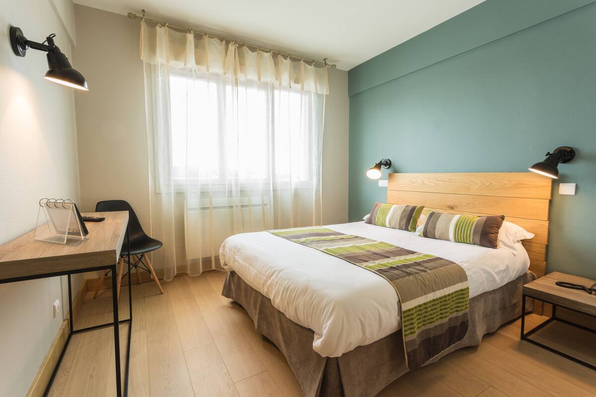 A neatly arranged bedroom with a bed, bedside table, desk, chair, and a window with curtains.