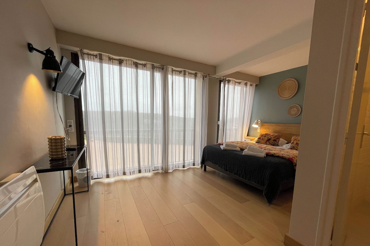 Un dormitorio con cama, televisor montado en la pared y ventana con cortinas traslúcidas.