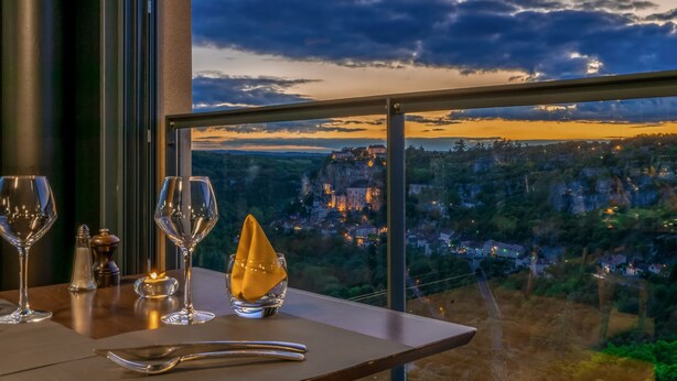 A table set with wine glasses, a salt shaker, a pepper shaker, and a napkin holder, overlooking a scenic view of a town nestled in a valley with a prominent building on a hill. - Le Belvedere (Rocamadour)