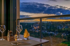 A table set with wine glasses, a salt shaker, a pepper shaker, and a napkin holder, overlooking a scenic view of a town nestled in a valley with a prominent building on a hill. - Le Belvedere (Rocamadour)
