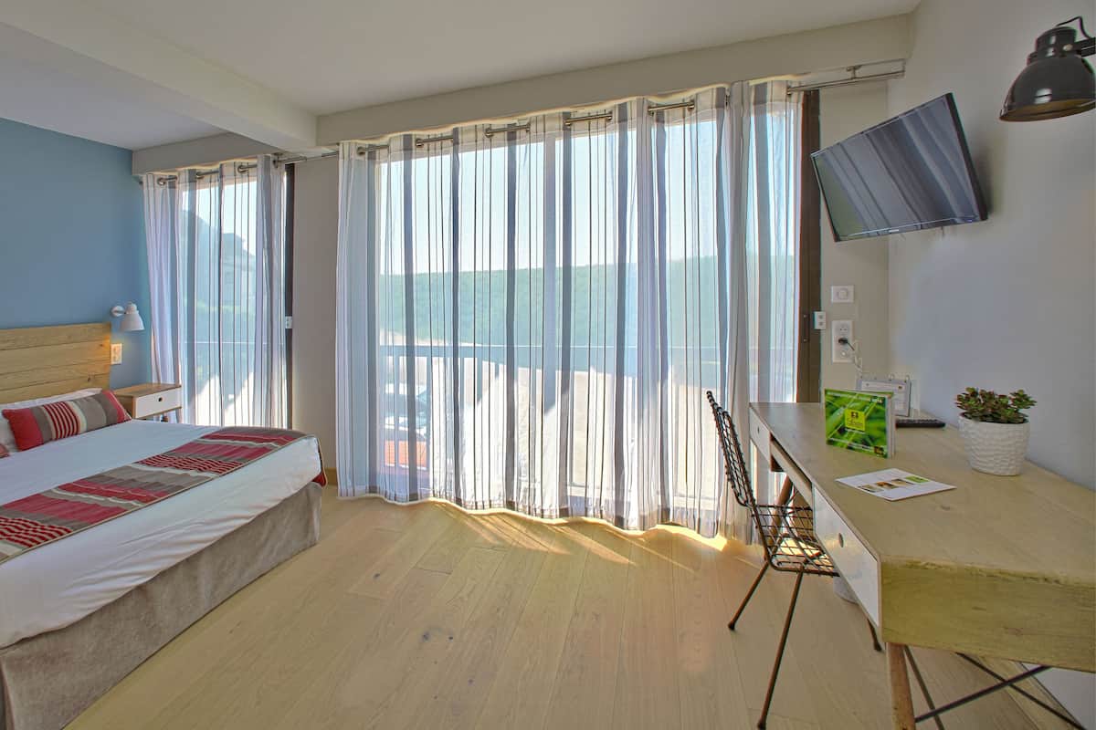 A hotel room with a bed, a desk with a chair, a TV, and a balcony with curtains.
