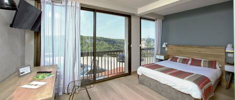Habitación de hotel con cama, escritorio, televisor y balcón con vistas al paisaje.