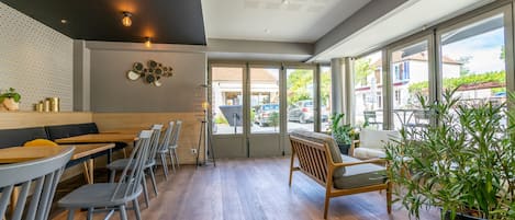 A modern cafe with wooden tables and chairs, a black and white patterned wall, and large windows offering a view of the street.
