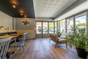 Un café moderno con mesas y sillas de madera, una pared con diseño blanco y negro y grandes ventanales que dan a la calle.