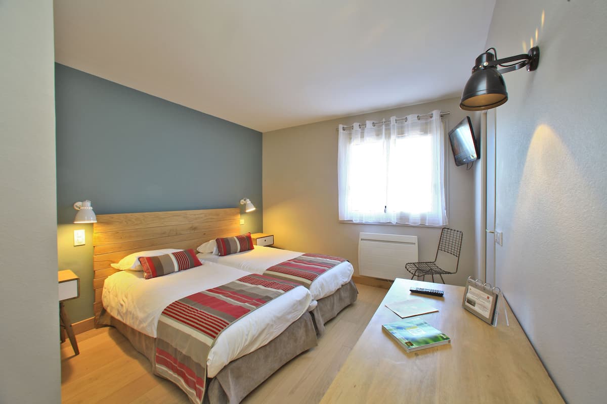 A bedroom with two beds, a desk, a chair, a TV, and a window with curtains.