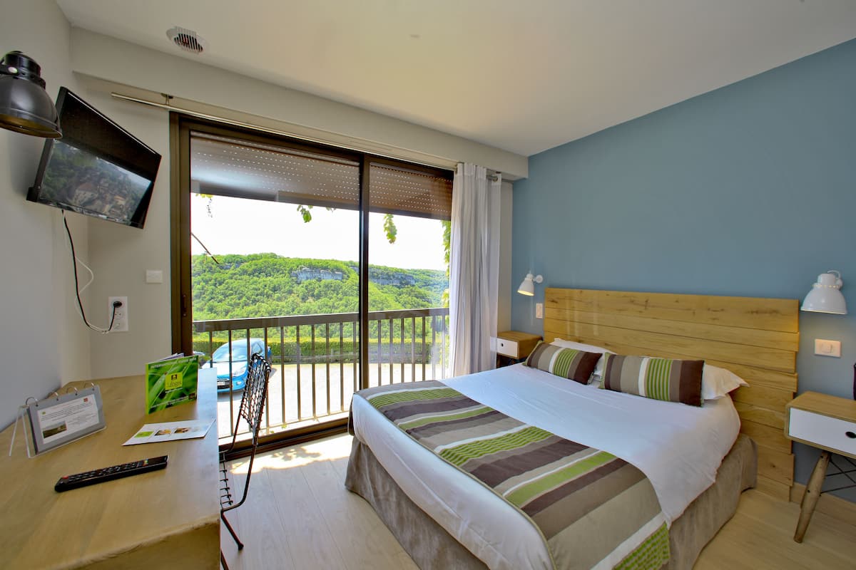 A hotel room with a bed, a desk with a computer, a TV mounted on the wall, and a balcony with a view of greenery.