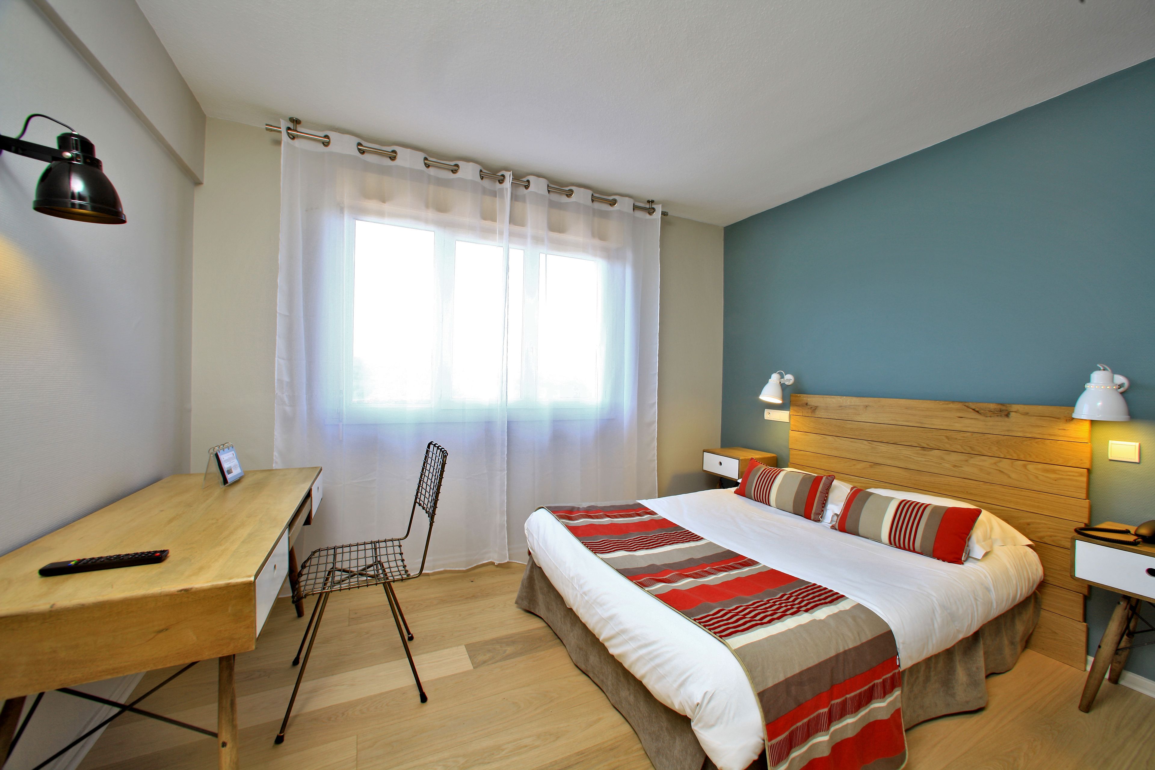 Habitación de hotel con un escritorio de madera, una cama con colcha a rayas, una silla y una ventana con cortinas traslúcidas.