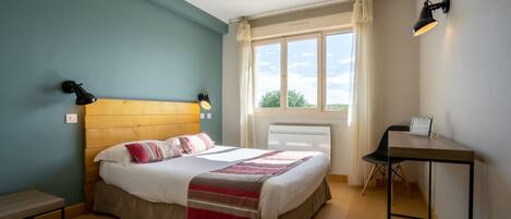 Ein Schlafzimmer mit einem hölzernen Kopfende, einem Bett mit weißer Bettwäsche und einer gestreiften Decke, einem Nachttisch, einem Sessel, einem Schreibtisch mit Lampe und einem Fenster mit Vorhängen.