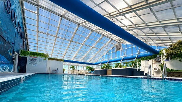 An indoor swimming pool with a glass ceiling, clear blue water, and a lifeguard warning sign. - Bolero Resort (Wildwood)