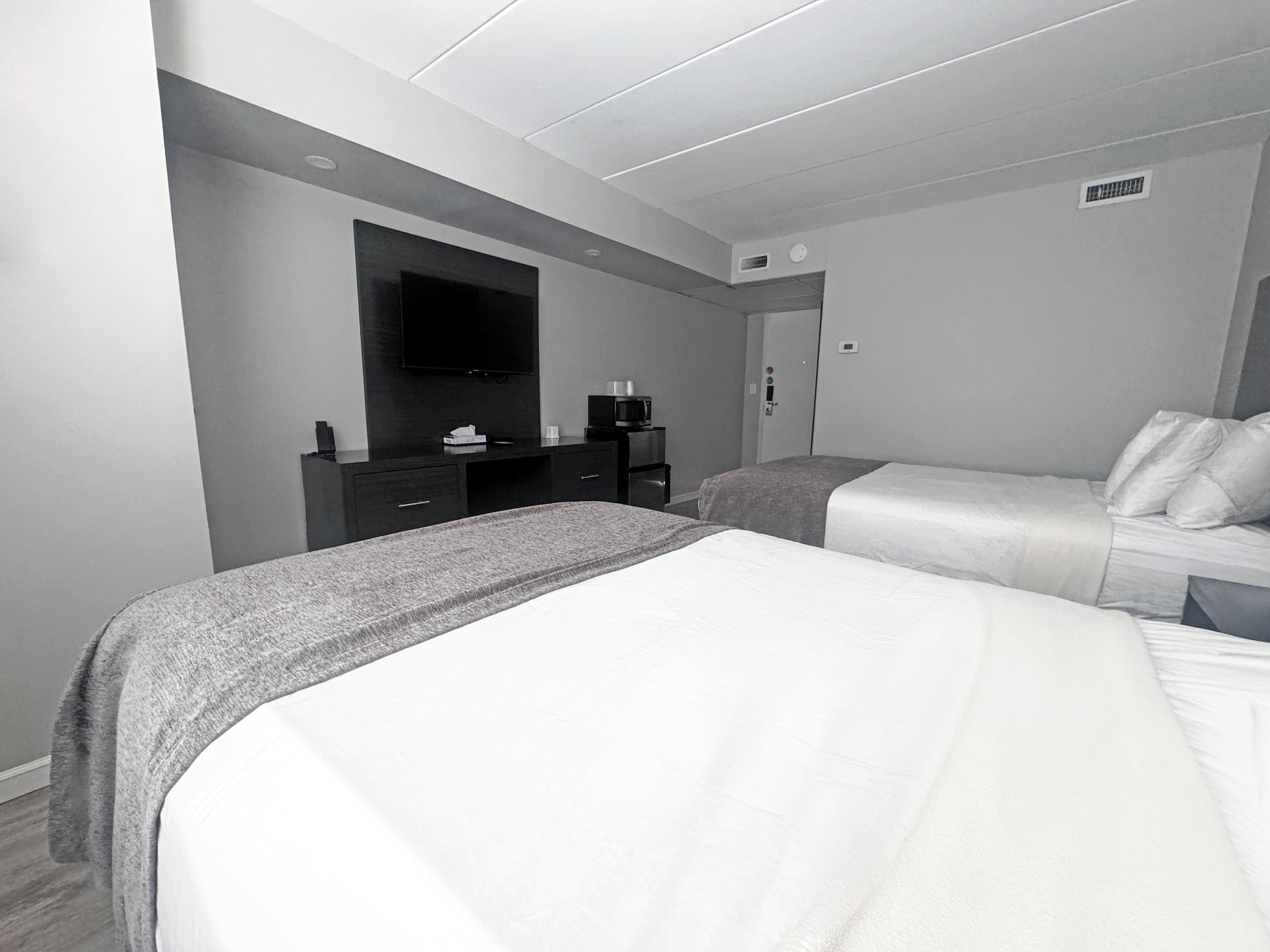 A modern hotel room with two beds, a flat-screen TV mounted on the wall, a small desk with a coffee maker, and a door with a handle.