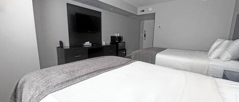 A modern hotel room with two beds, a flat-screen TV mounted on the wall, a small desk with a coffee maker, and a door with a handle.