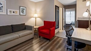 A modern hotel room with a wooden floor, a red armchair, a grey sofa, a desk with a chair, and a lamp. - Country Inn & Suites by Radisson, Salisbury, MD (Salisbury)