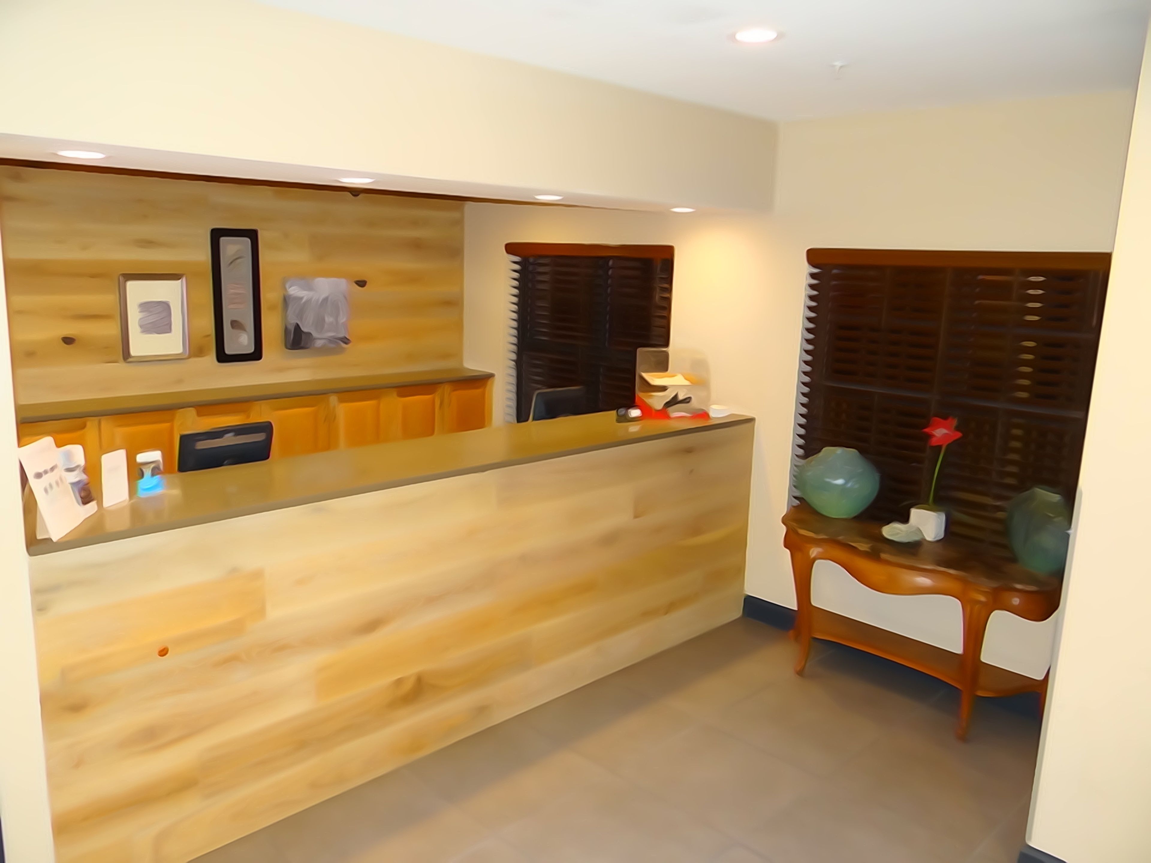 A reception desk with a wooden finish, a small table with a vase and a red flower, and a window with blinds.