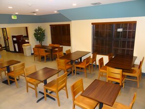 A restaurant with multiple wooden tables and chairs, a potted plant, and a window with blinds.
