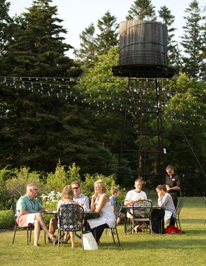 An outdoor gathering with people seated at tables, surrounded by greenery and a water tower in the background.