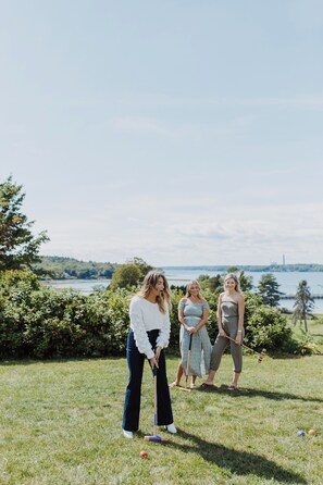 Three people playing croquet in an outdoor setting with a view of a body of water and trees in the background.
