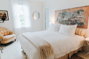 A spacious bedroom with a large bed, a round mirror, a painting, and a chair.