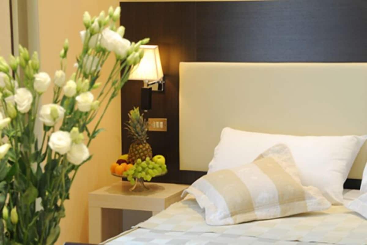 A hotel room with a bed, white pillows, a nightstand with a fruit bowl, a lamp, and a wall-mounted light switch.