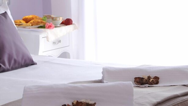 A neatly made bed with white linens and two pillows, a bedside table with a bowl of fruit and a cup, and a window with curtains. - Hotel I Due Cigni (Montepulciano)
