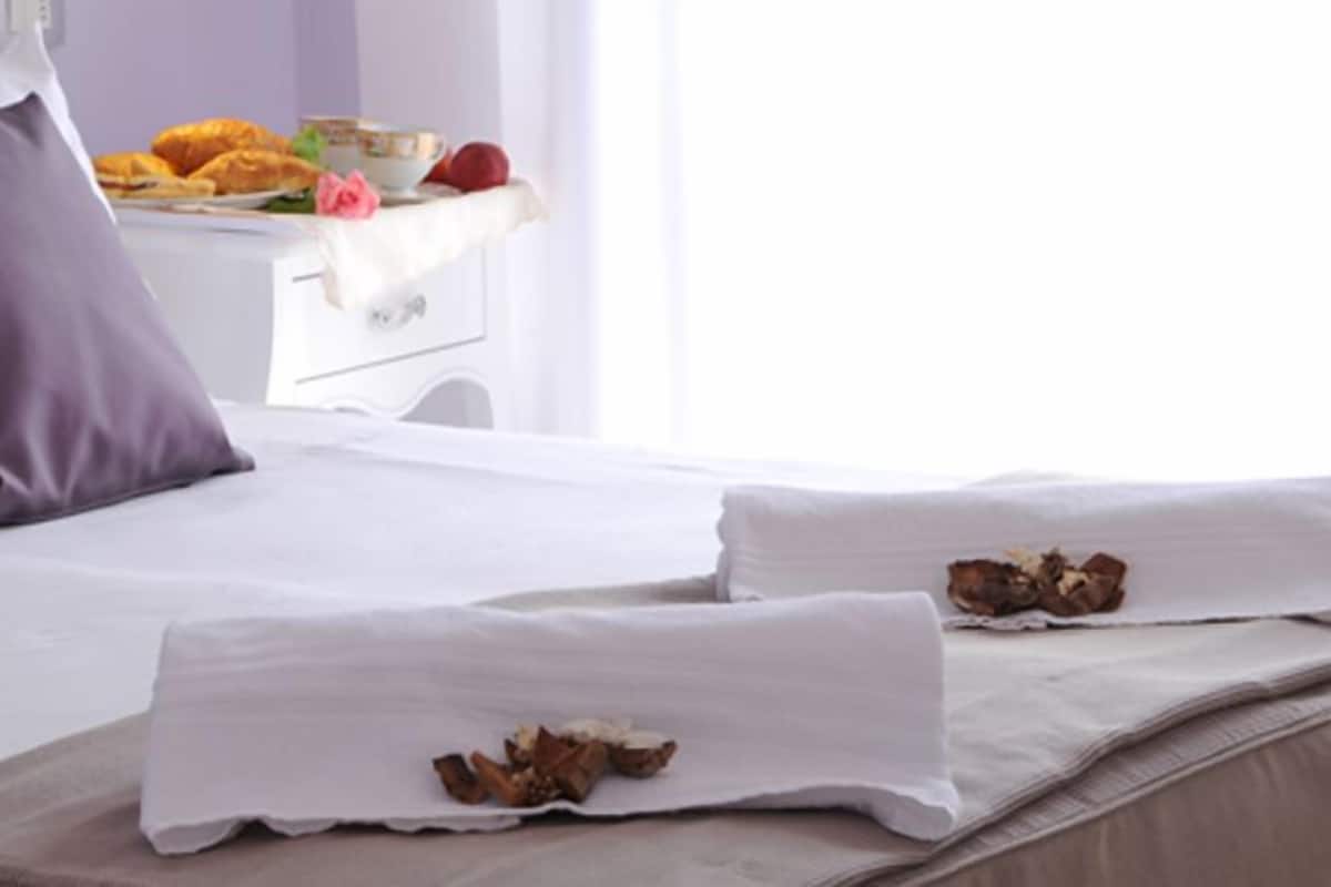 A neatly made bed with white linens and two pillows, a bedside table with a bowl of fruit and a cup, and a window with curtains.