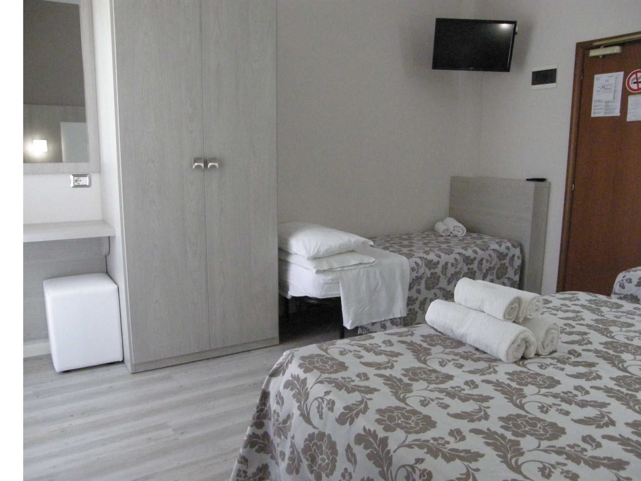 Ein Hotelzimmer mit zwei Betten, einem Kleiderschrank, einem an der Wand befestigten Fernseher und einer kleinen weißen Kommode.