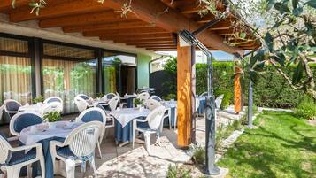Een terras buiten met witte stoelen en tafels, overdekt met een houten pergola, omringd door groen.
