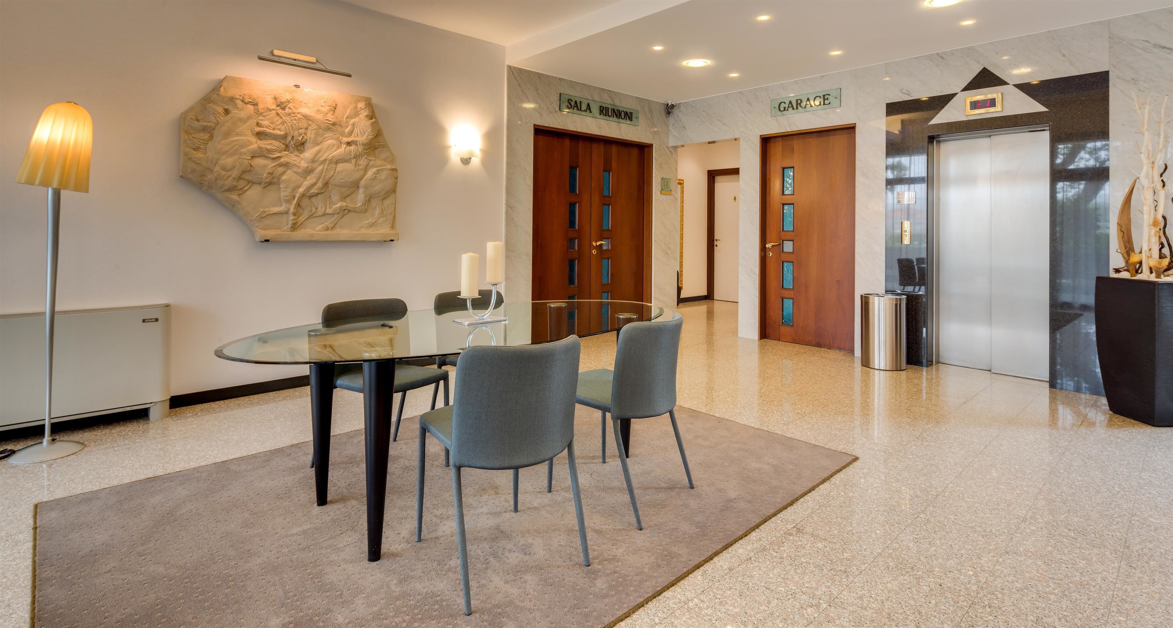 A modern lobby with a glass table, two chairs, a sculpture, a reception desk, and a sign indicating the location of the Sala Riunioni and Garage.
