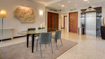 A modern lobby with a glass table, two chairs, a sculpture, a reception desk, and a sign indicating the location of the Sala Riunioni and Garage.