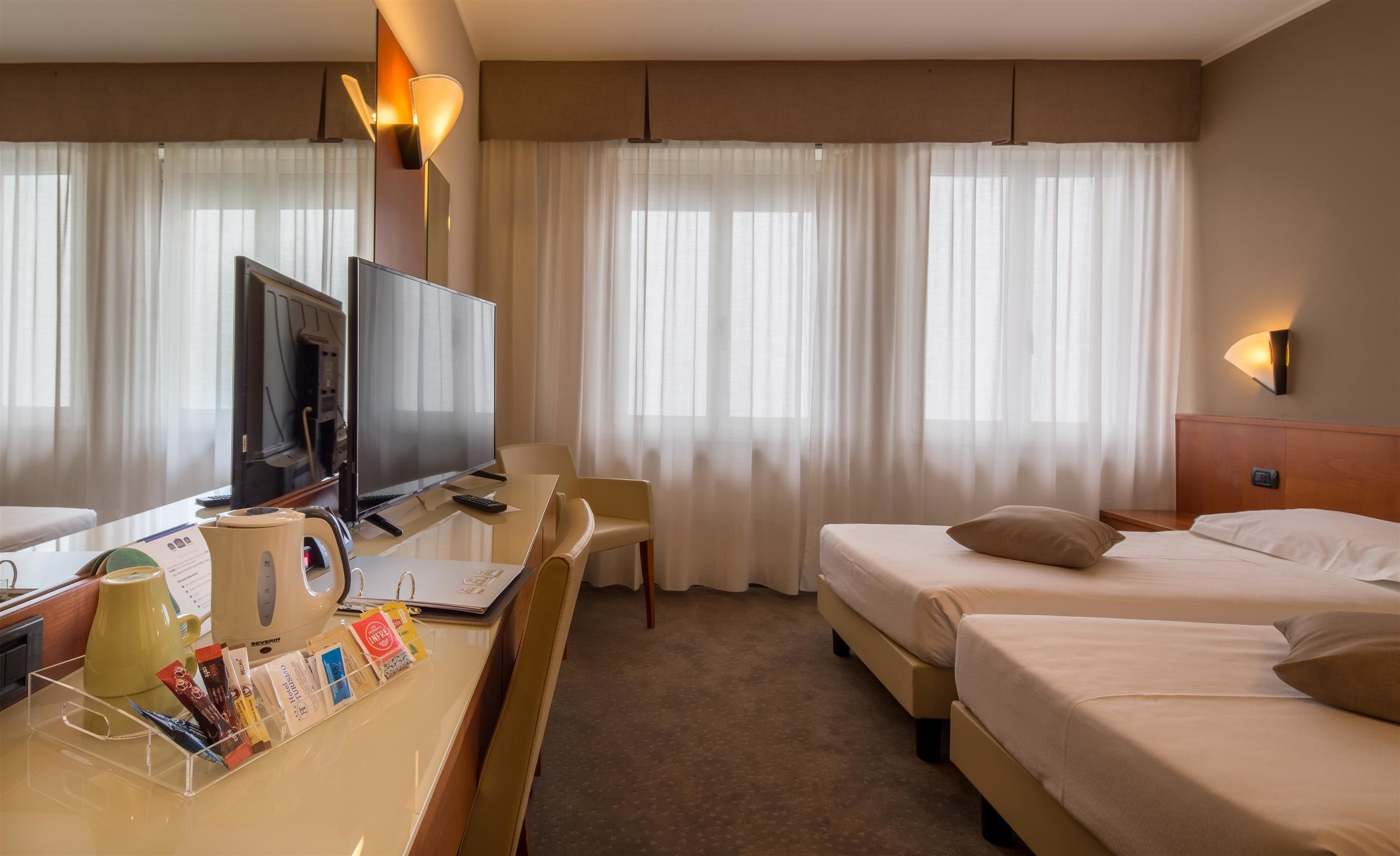 A hotel room with a desk, TV, two beds, and large windows with sheer curtains.