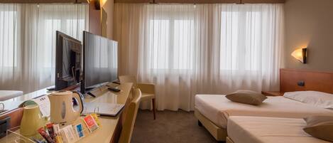 A hotel room with a desk, TV, two beds, and large windows with sheer curtains.