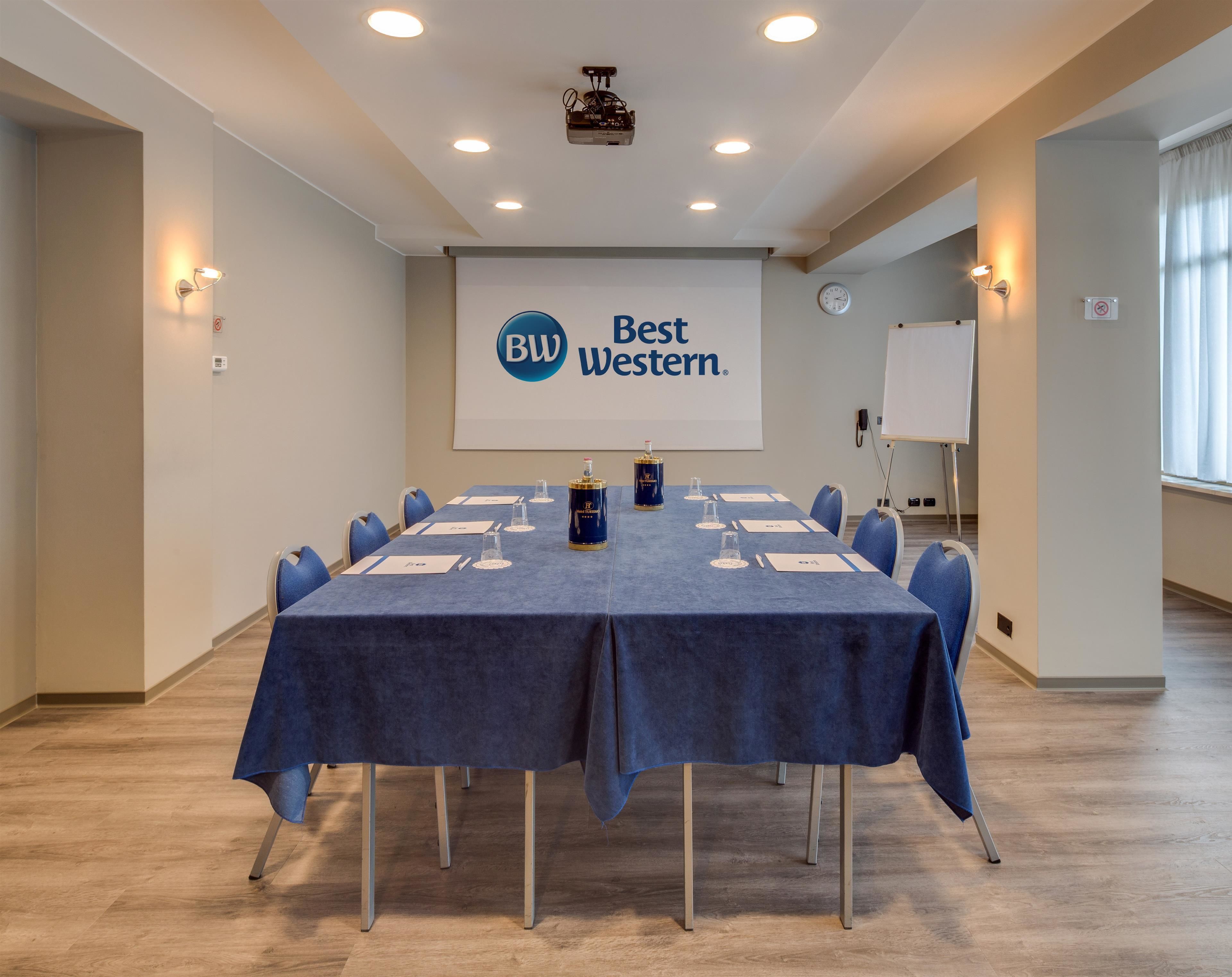 Un salón de conferencias con una larga mesa, sillas, un proyector y una pantalla que muestra la marca Best Western.