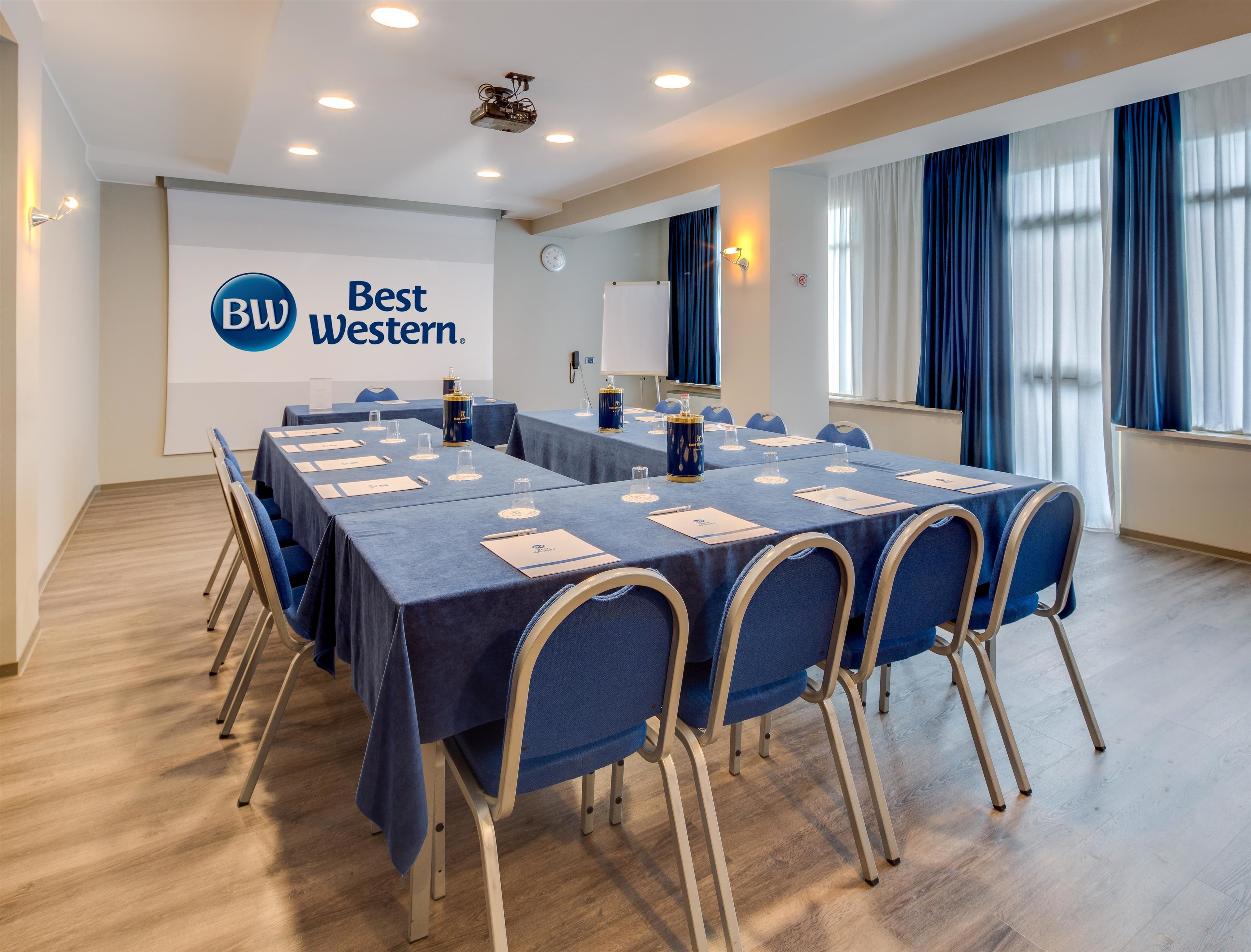Een congreszaal met een lange tafel, stoelen, een projecteur en een scherm waarop de branding van Best Western staat.