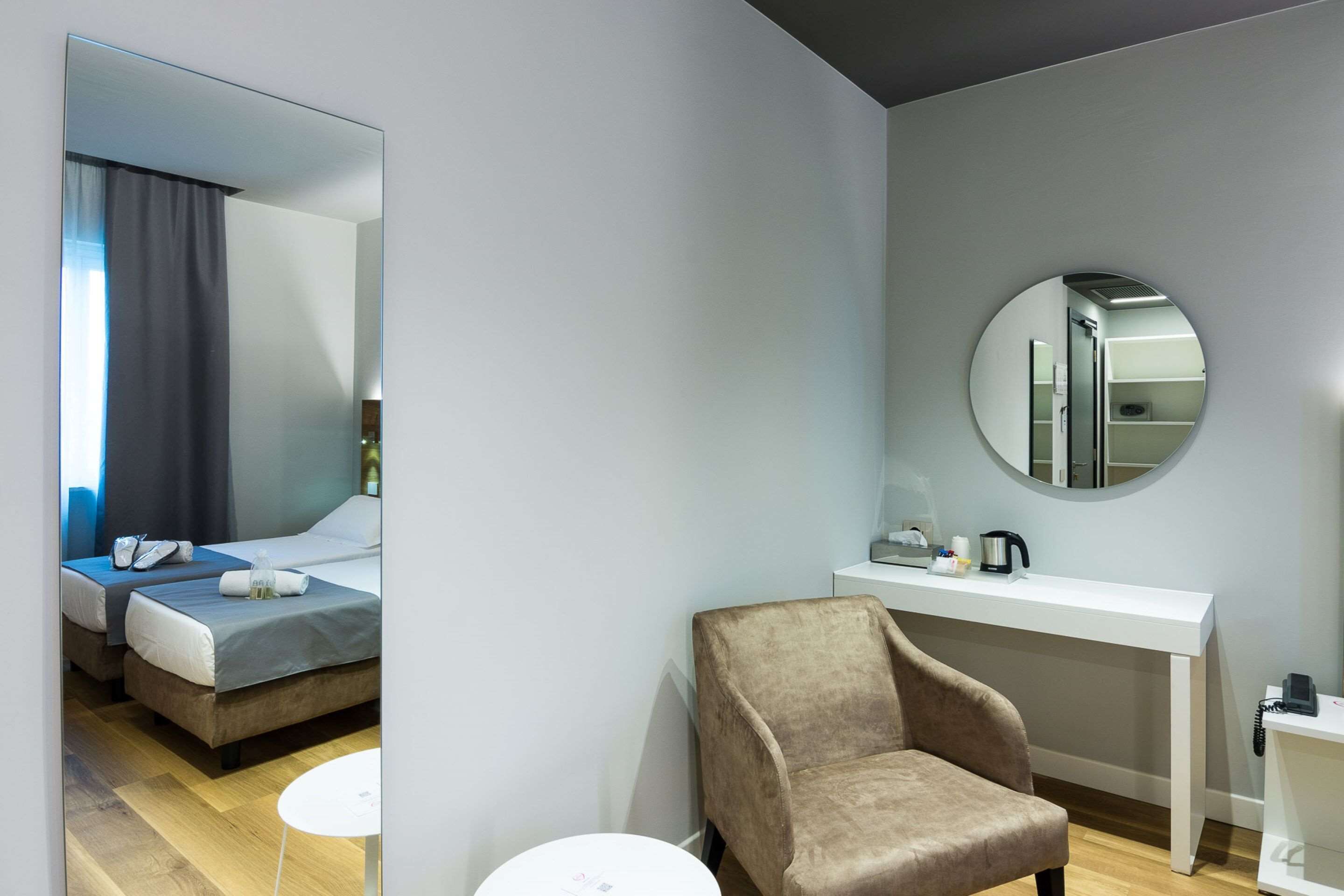 Una habitación de hotel moderna con un espejo grande, un espejo redondo, una cama, una silla, una mesita pequeña y una consola blanca con una tetera y una cafetera.