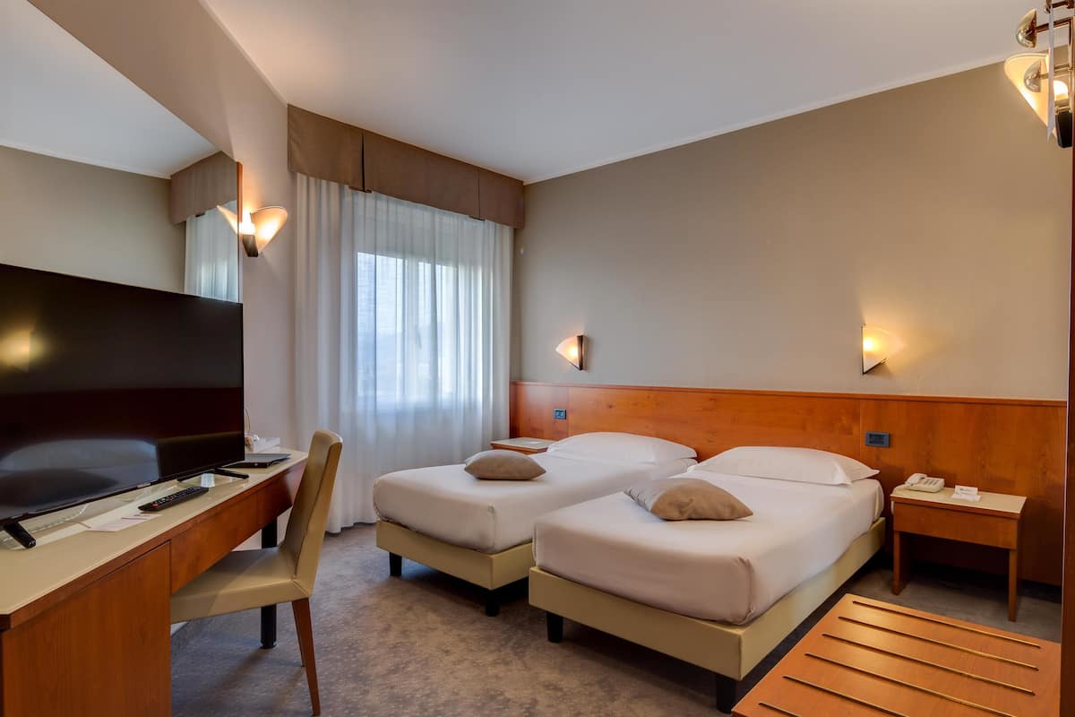 A hotel room with two beds, a desk with a TV, a chair, a bedside table, and a wooden side table.
