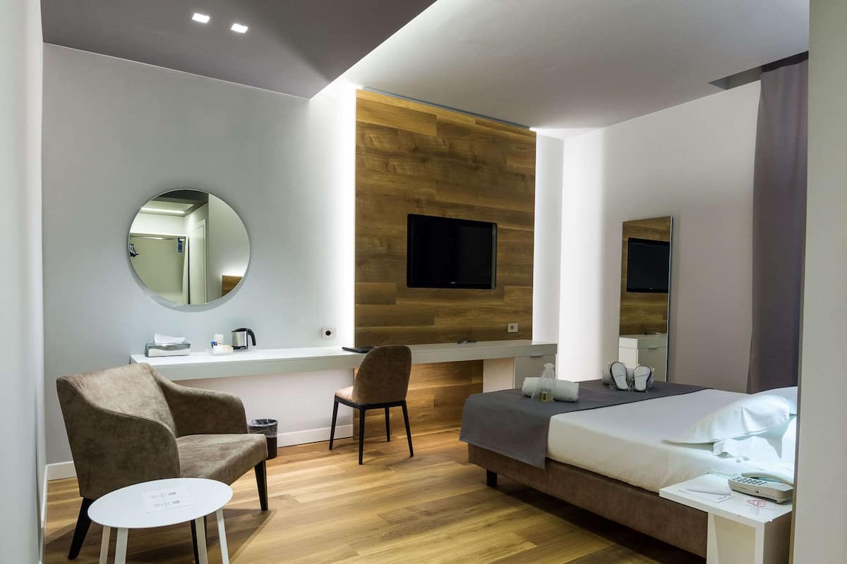 A modern hotel room with a wooden accent wall, a flat-screen TV, a bed, a chair, a small table, and a mirror.