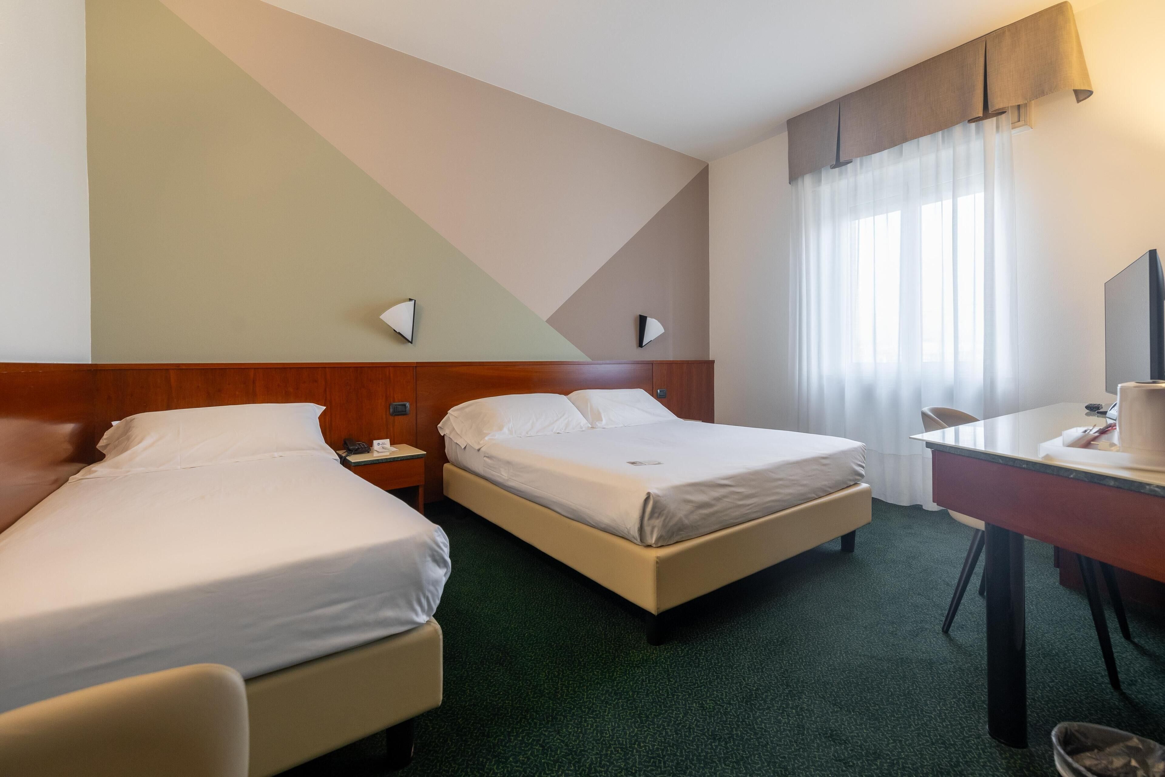 A hotel room with two beds, a desk, a chair, a TV, and a window with curtains.
