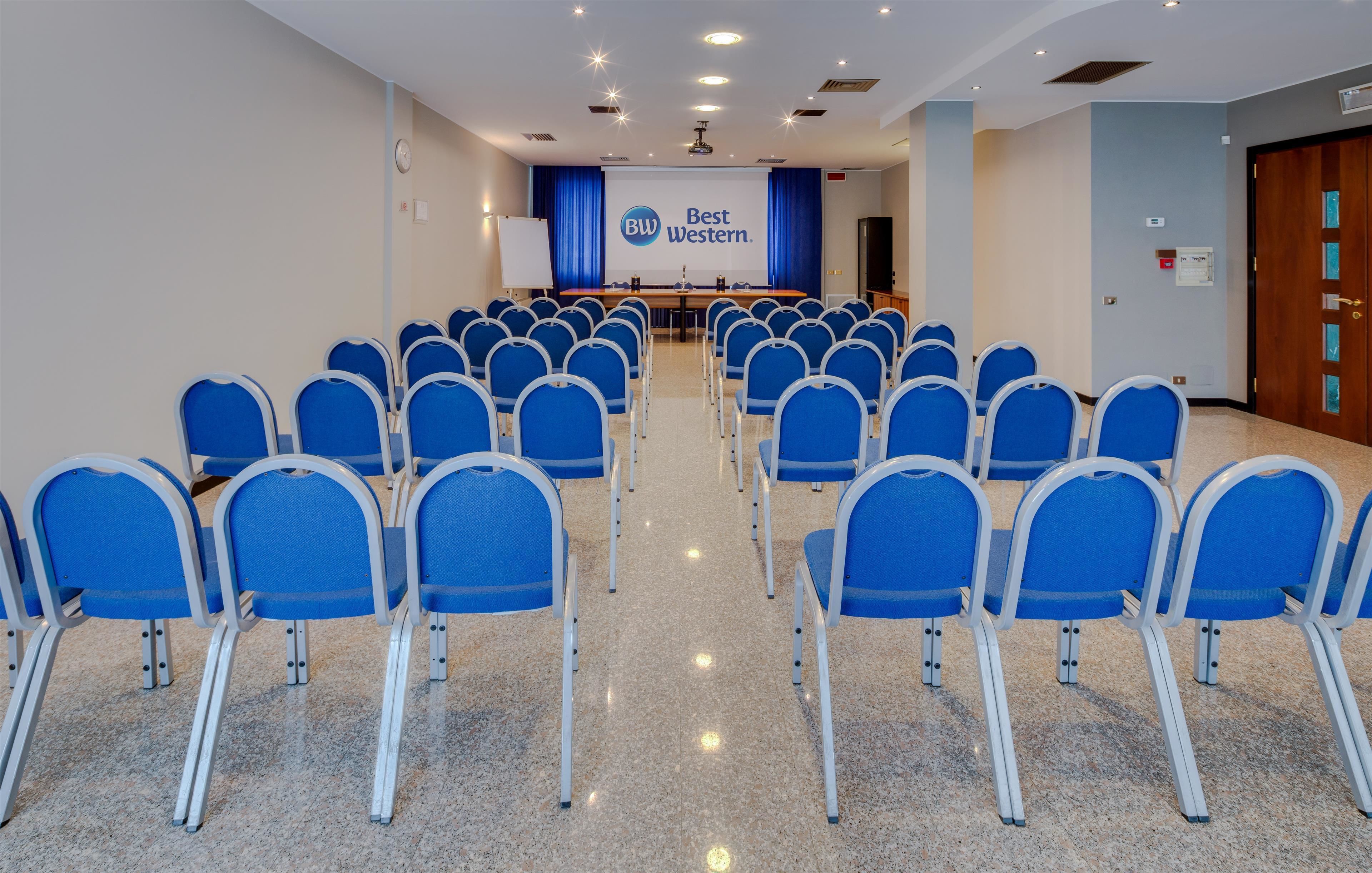 A conference room with rows of blue chairs, a stage with a screen displaying 'Best Western', and a door on the right.