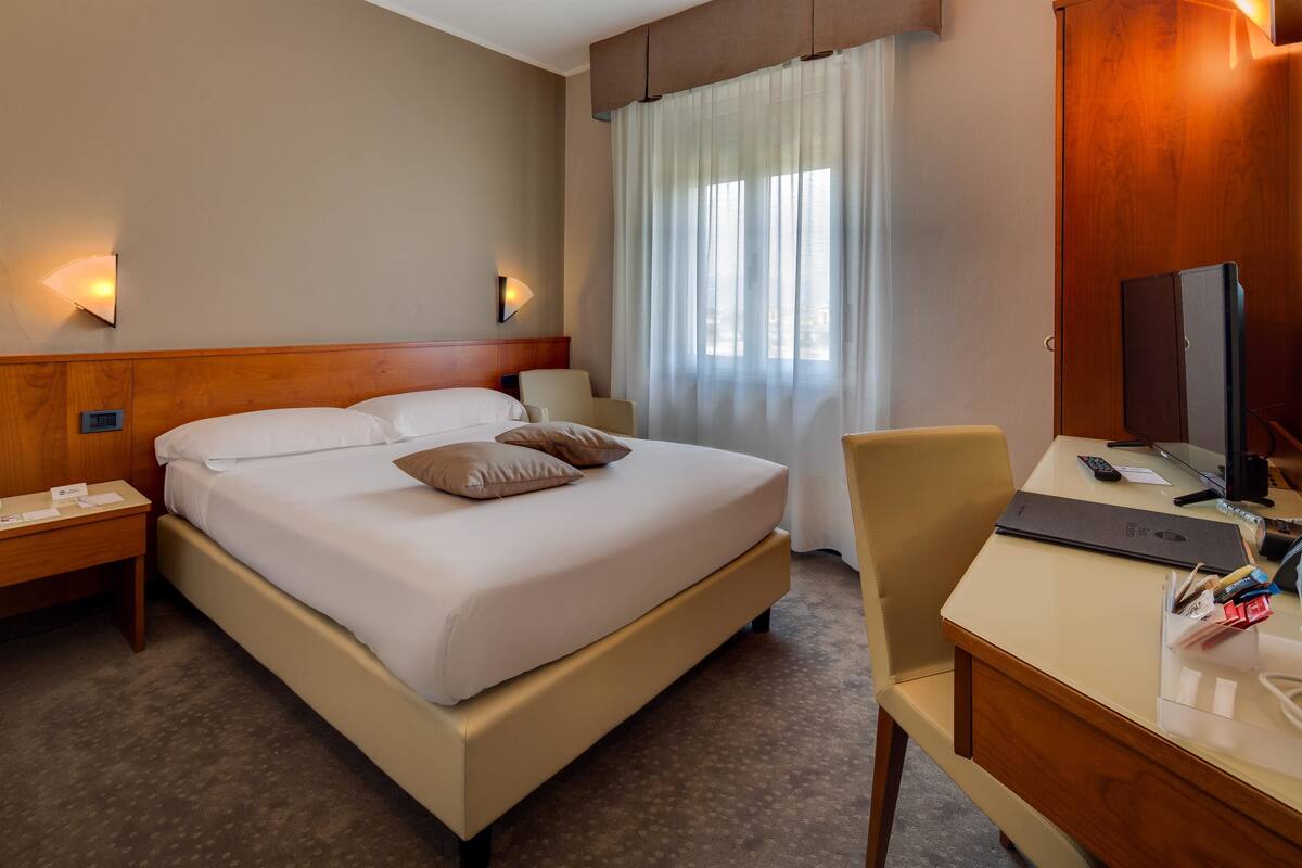 A hotel room with a bed, bedside table, chair, desk, and a TV.
