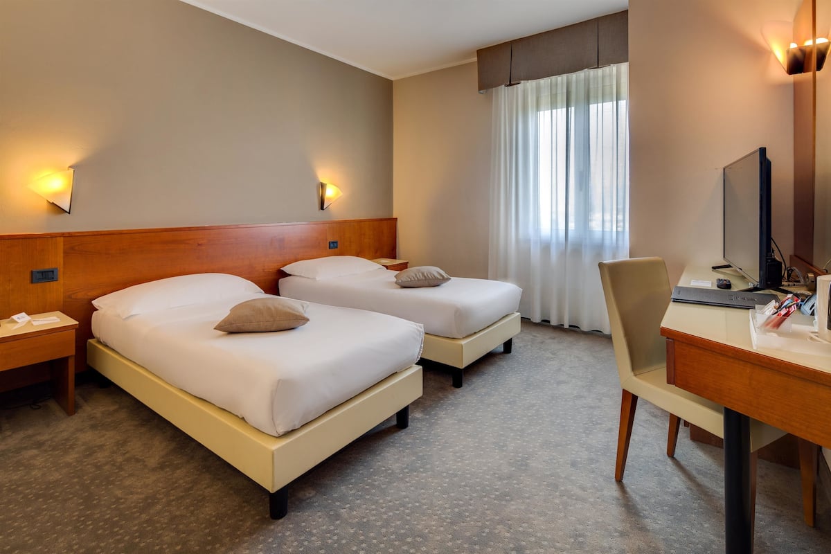 A hotel room with two beds, a desk, a chair, and a large window with curtains.