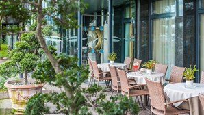 An outdoor seating area with wicker chairs and round tables, surrounded by glass walls and a large decorative clock. - Best Western Hotel Turismo (San Martino Buon Albergo)