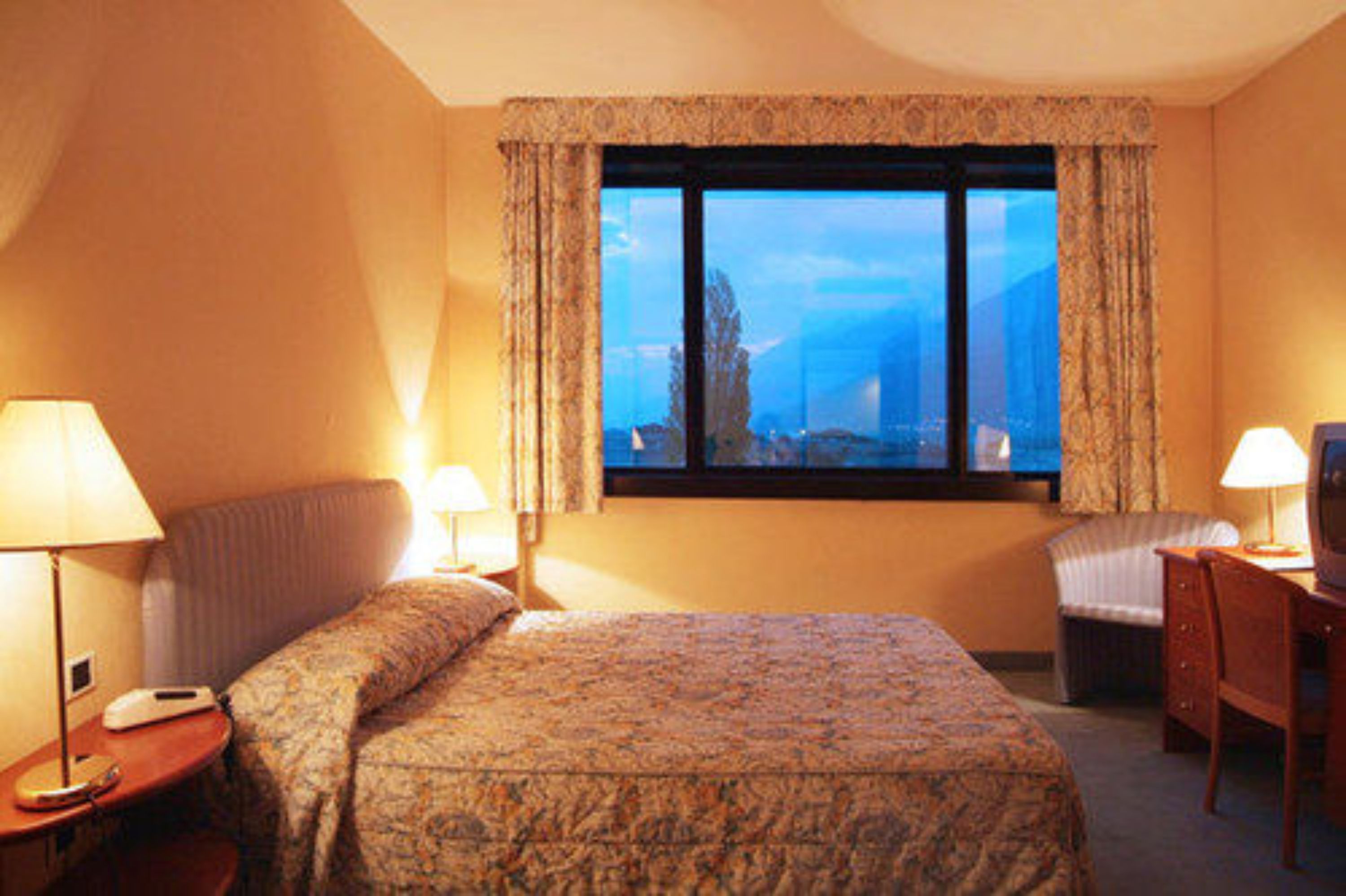 Habitación de hotel con una cama, dos mesitas de noche con lámparas, un escritorio con silla y un ventanal que ofrece vistas a las montañas.
