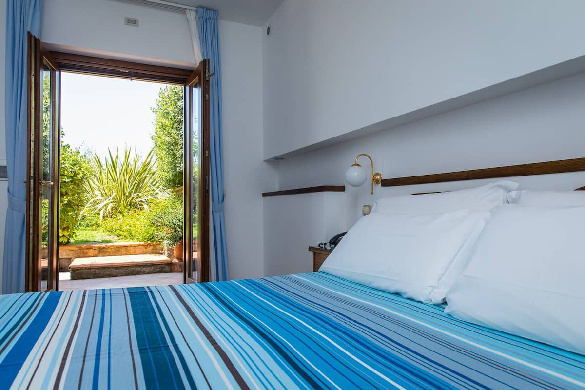 Una habitación de hotel con una cama, una ventana con cortinas, una lámpara de noche y vistas al exterior con vegetación.