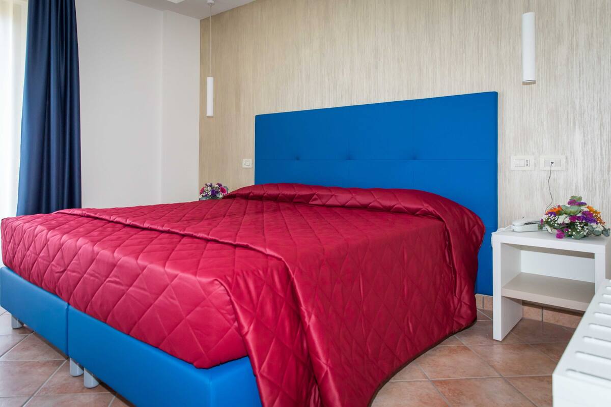 Una habitación de hotel con cabecera azul, colcha de cama acolchada roja y una mesita de noche blanca con un florero.