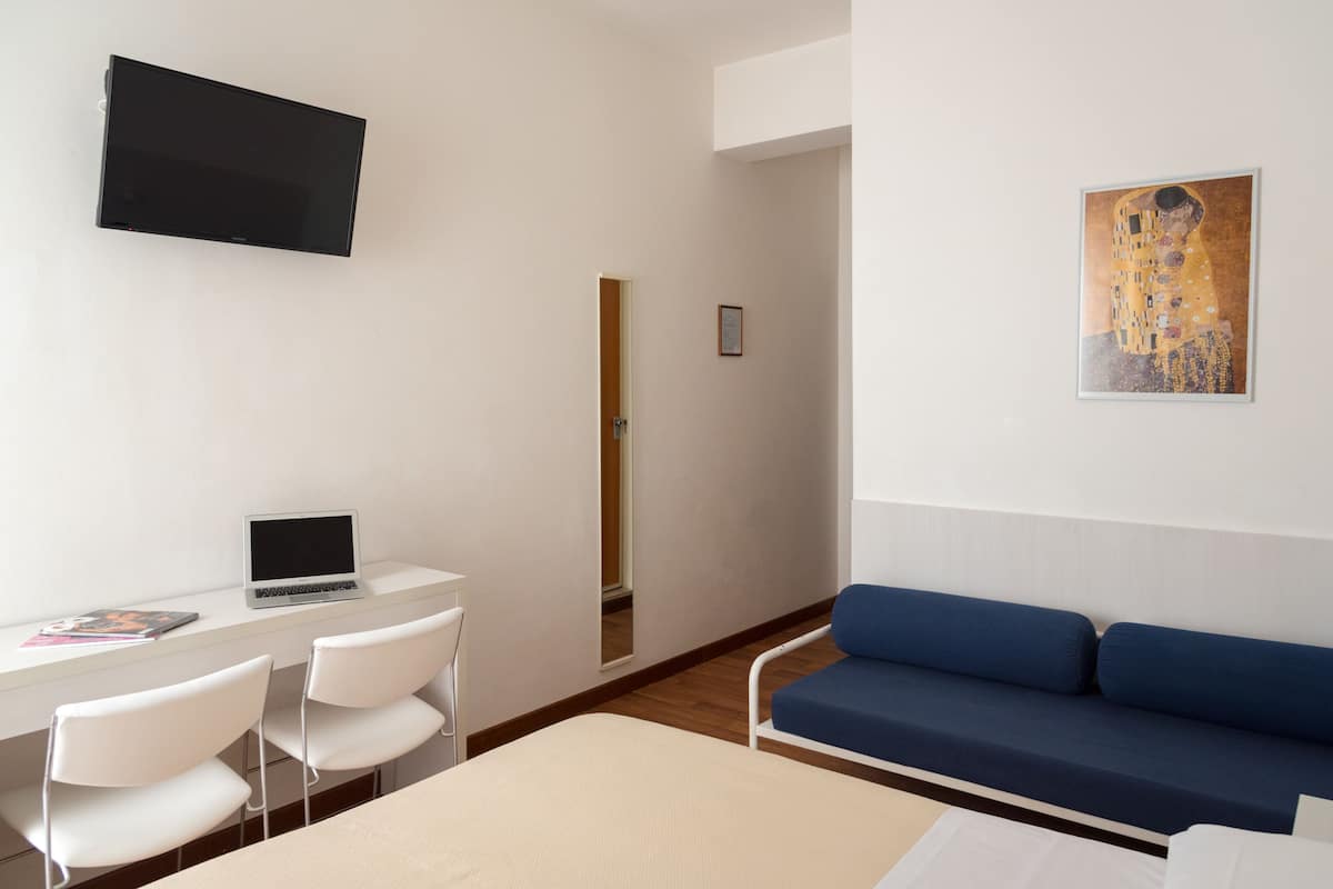A modern hotel room with a flat-screen TV mounted on the wall, a desk with a laptop, two white chairs, a blue sofa, and a framed artwork on the wall.