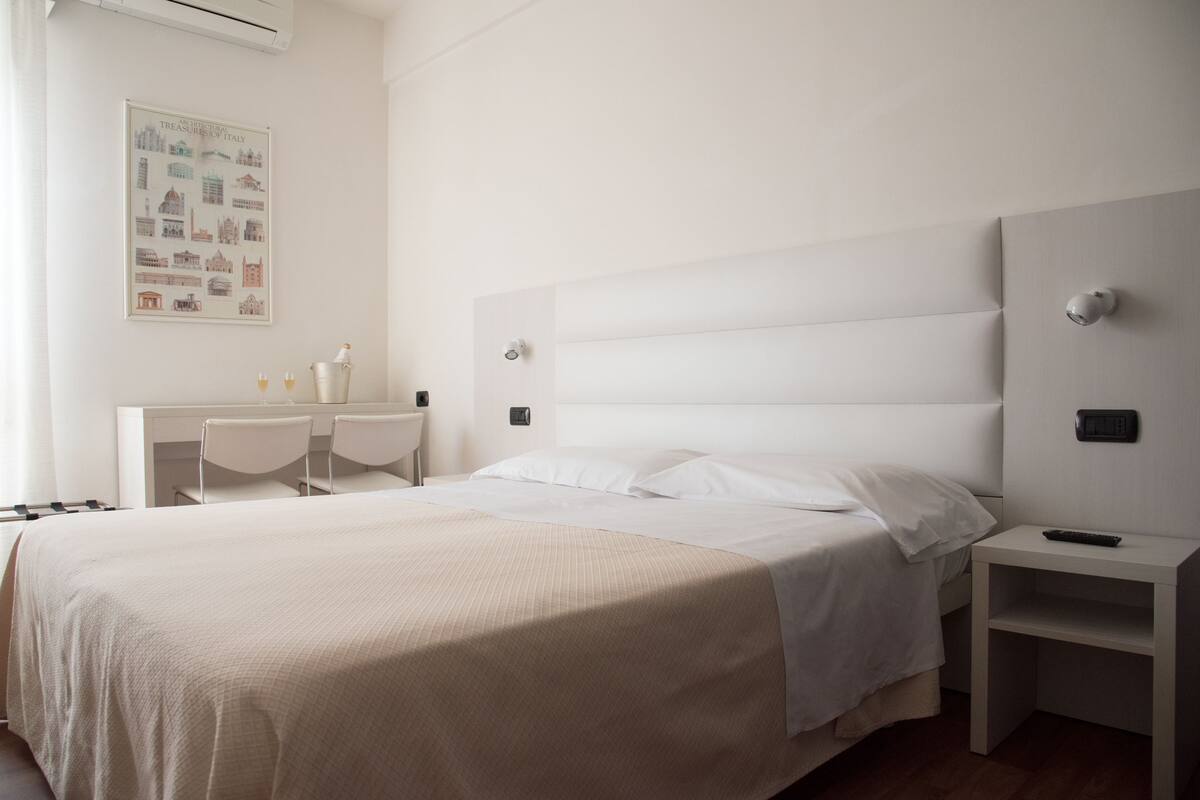 A minimalist hotel room with a large bed, a desk with two chairs, a small table, and a wall-mounted air conditioner.