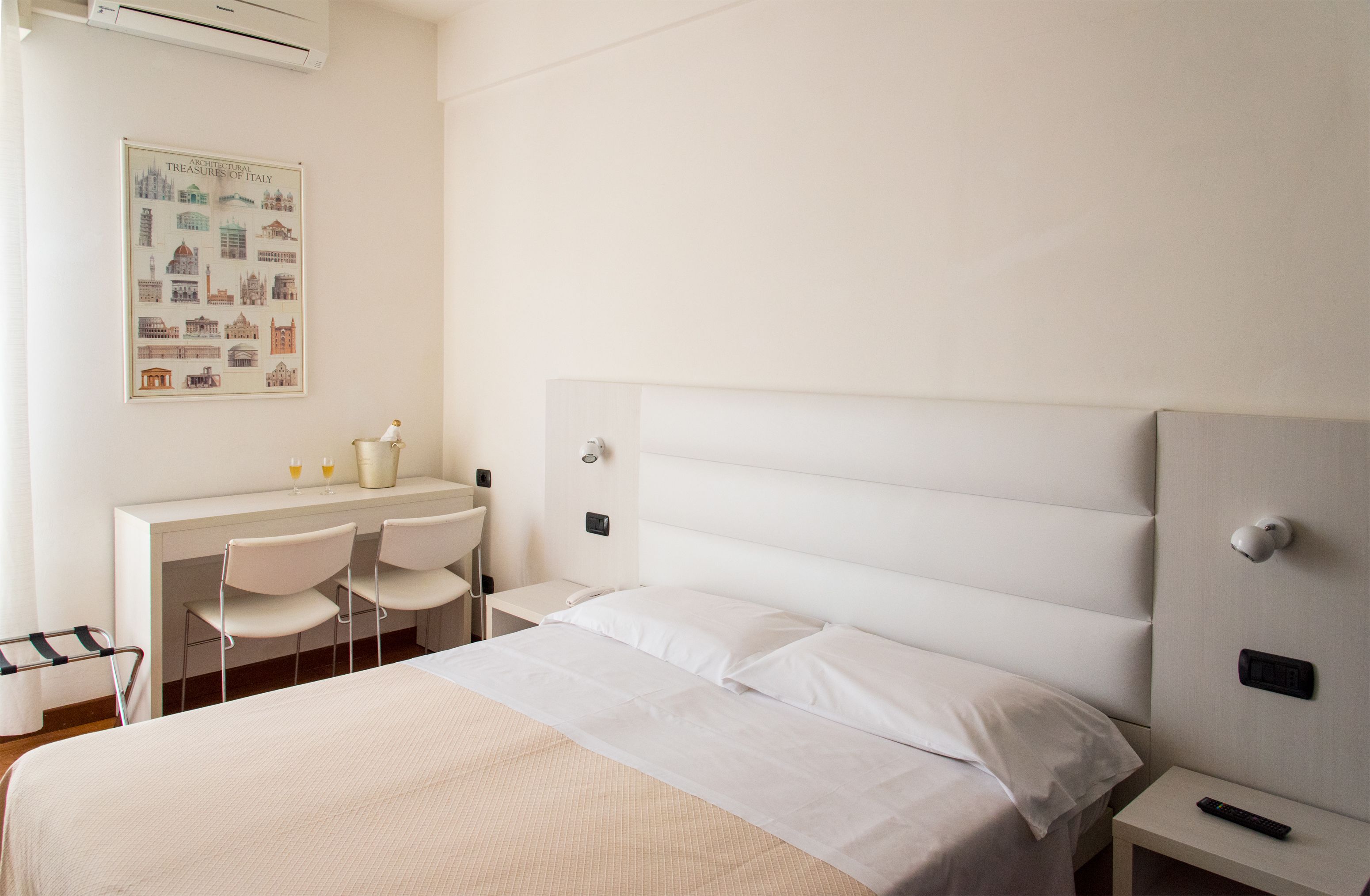 A hotel room with a bed, a desk with two chairs, a small table, and a poster on the wall.